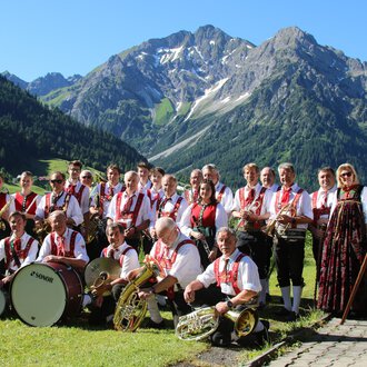 A brass band in traditional attire stands in a picturesque mountain landscape. In the background, there are tall mountains and a blue sky. | © Musikverein d´Hirschegger Kleinwalsertal