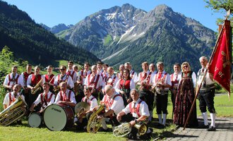 A brass band in traditional attire stands in a picturesque mountain landscape. In the background, there are tall mountains and a blue sky. | © Musikverein d´Hirschegger Kleinwalsertal