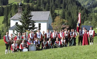 A traditional costumes band stands on a meadow in front of a small church. In the background, green hills and trees can be seen. | © Trachtenkapelle Harmonie Mittelberg | Jockl Metzger