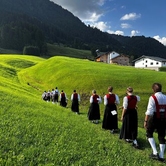 Eine traditionelle Blaskapelle in Trachten steht auf einer Wiese vor einem rustikalen Holzhaus. Die Musiker halten ihre Instrumente und lächeln in die Kamera, mit Bergen im Hintergrund. | © Trachtenkapelle Riezlern