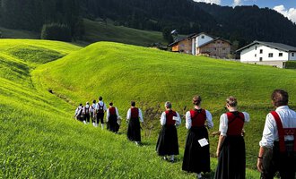 Eine traditionelle Blaskapelle in Trachten steht auf einer Wiese vor einem rustikalen Holzhaus. Die Musiker halten ihre Instrumente und lächeln in die Kamera, mit Bergen im Hintergrund. | © Trachtenkapelle Riezlern