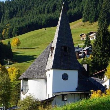 Eine malerische Kirche mit einem hohen, spitzen Turm inmitten einer grünen Landschaft. Im Hintergrund sind sanfte Hügel und einige traditionelle Häuser zu sehen. | © Ev. Kirche Kleinwalsertal