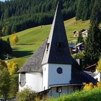 A picturesque church with a tall, pointed tower amidst a green landscape. In the background, gentle hills and some traditional houses are visible. | © Ev. Kirche Kleinwalsertal