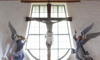 A small church room with a cross and angels on the walls. The altar is decorated with flowers and surrounded by candles. | © Ev. Kirche Kleinwalsertal