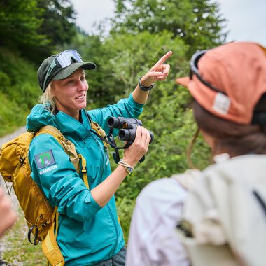 A woman with a camera is explaining something to a group of people outdoors. In the background, there are trees and a hilly landscape visible. | © Kleinwalsertal Tourismus eGen | Oliver Farys