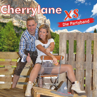 A couple is posing on a rustic wooden bench in front of a beautiful mountain landscape. They are wearing traditional clothing and smiling happily at the camera.
