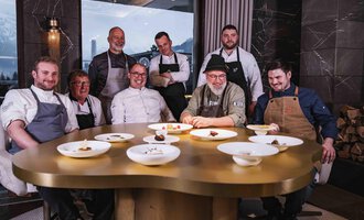 A group of chefs is sitting at a round table with several elegant plates. In the background, there are large windows with a view of the mountains. | © Frank Simon