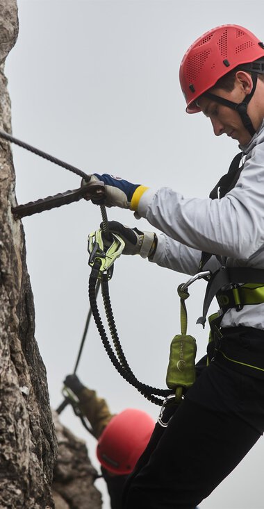 A climber is secured to a rock wall and is focusing on his technique. In the background, another climber can be seen, while a misty atmosphere prevails. | © Bergschule Kleinwalsertal | Oliver Farys