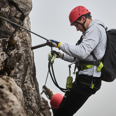 A climber is secured to a rock wall and is focusing on his technique. In the background, another climber can be seen, while a misty atmosphere prevails. | © Bergschule Kleinwalsertal | Oliver Farys