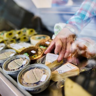 Ein Verkaufsstand mit verschiedenen Käsesorten. Eine Hand greift nach einem Käseprodukt in der Auslage. | © Kleinwalsertal Tourismus | Fotograf Oliver Farys
