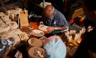 Ein älterer Mann schnitzt Holzfiguren an einem Marktstand. Um ihn herum liegen zahlreiche Holzplatten mit verschiedenen Motiven. | © Kleinwalsertal Tourismus | Fotograf Oliver Farys