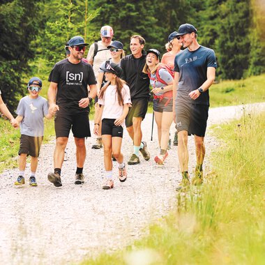 Eine Gruppe von Menschen wandert auf einem Kiesweg durch eine grüne Landschaft. Sie tragen Freizeitkleidung und scheinen viel Spaß zu haben. | © Kleinwalsertal Tourismus | Oliver Farys