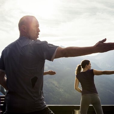 Eine Gruppe von Menschen übt in der Natur Tai Chi. Im Hintergrund sind majestätische Berge und ein leicht bewölkter Himmel zu sehen. | © Kleinwalsertal Tourismus | Oliver Farys