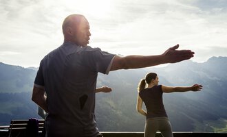 Eine Gruppe von Menschen übt in der Natur Tai Chi. Im Hintergrund sind majestätische Berge und ein leicht bewölkter Himmel zu sehen. | © Kleinwalsertal Tourismus | Oliver Farys