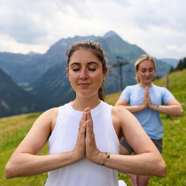 Zwei Frauen praktizieren Yoga in den Bergen. Die Umgebung ist grün und beeindruckend, mit hohen Bergen und einer bewölkten Himmel. | © Kleinwalsertal Tourismus | Oliver Farys