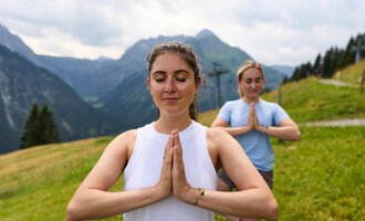 Zwei Frauen praktizieren Yoga in den Bergen. Die Umgebung ist grün und beeindruckend, mit hohen Bergen und einer bewölkten Himmel. | © Kleinwalsertal Tourismus | Oliver Farys