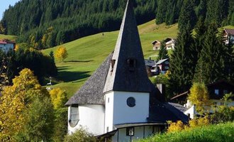 Ein traditionelles Gebäude mit einem hohen spitzen Dach steht in einer malerischen Landschaft. Umgeben von grünen Hügeln und bunten Bäumen wirkt die Szene friedlich und einladend. | © Evangelische Kreuzkirche Hirschegg