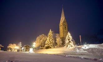 A snowy landscape at night with a church and illuminated trees. The sky is dark blue and the road is visible. | © Kleinwalsertal Tourismus | Dominik Berchtold