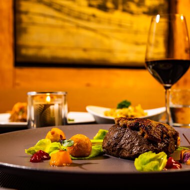 An elegant dinner featuring a juicy steak on a plate, surrounded by colorful vegetables. In the background, wine glasses and additional dishes are visible, all in warm lighting.