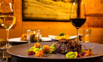 An elegant dinner featuring a juicy steak on a plate, surrounded by colorful vegetables. In the background, wine glasses and additional dishes are visible, all in warm lighting.