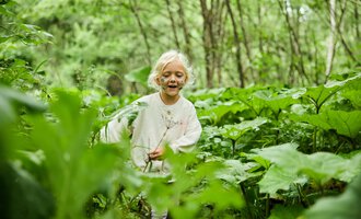 Ein Mädchen mit blonden Haaren spielt fröhlich in einem grünen Wald. Umgeben von großen Pflanzen strahlt sie Freude und Unbeschwertheit aus. | © Kleinwalsertal Tourismus | Oliver Farys