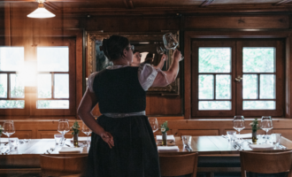 Eine Kellnerin in traditioneller Tracht steht mit einem Glas in der Hand in einem gemütlichen Restaurant. Der Holzraum ist mit Tischen und stilvollen Fenstern ausgestattet. | © Winzer zu Tisch | Naturhotel Chesa Valisa
