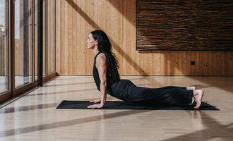 A bright yoga room with wooden paneling and large windows. Three people are practicing yoga and looking outside. | © Naturhotel Chesa Valisa