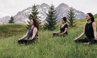 Ein heller Yogaraum mit Holzverkleidung und großen Fenstern. Drei Personen praktizieren Yoga und blicken nach draußen. | © Naturhotel Chesa Valisa