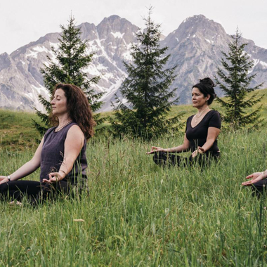 A bright yoga room with wooden paneling and large windows. Three people are practicing yoga and looking outside. | © Naturhotel Chesa Valisa
