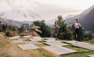 Two women are practicing yoga on wooden platforms in a green meadow. In the background, mountains and a picturesque landscape can be seen.