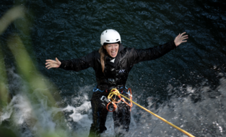 Eine Person klettert begeistert an einem Seil über Wasser. Sie trägt einen Helm und ist in einem Schutzanzug gekleidet.