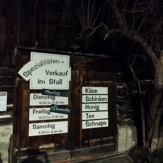 © Abler Spezialitätenverkauf im Stall | N. Lughammer A wooden building with a sign for specialty sales in the barn. The signage indicates opening hours and offered products such as cheese and honey. | © Abler Spezialitätenverkauf im Stall | N. Lughammer