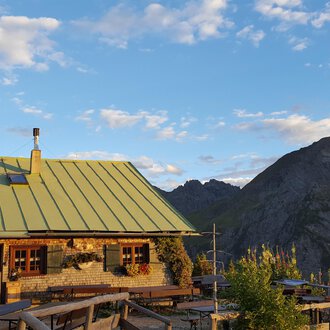 A cozy mountain hut with a green roof in the Alps. In the background, majestic mountains stretch out under a blue sky. | © Alpe Kuhgehren | Lisa Hiesinger