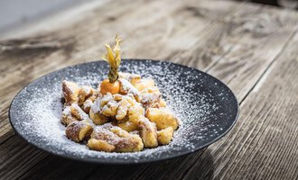 Ein schwarzes Teller voller gebackener Stücke, bestreut mit Puderzucker. Ein kleines Stück Obst schmückt die Präsentation. | © Alpenhotel DAS KÜREN