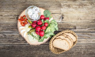 Ein rustikales Holzbrett mit frischem Gemüse, Radieschen und eingelegten Gurken. Daneben liegt ein Korb mit Scheiben Vollkornbrot. | © Alpenhotel DAS KÜREN