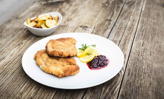 Ein Teller mit zwei panierten Schnitzeln, serviert mit Zitronenscheiben und einer Portion Preiselbeeren. Im Hintergrund befindet sich eine Schüssel mit Pommes. | © Alpenhotel DAS KÜREN