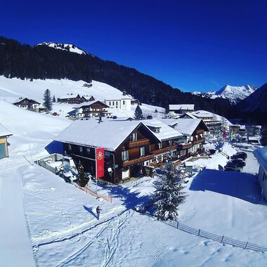 Eine schneebedeckte Landschaft mit gemütlichen Holzhaus und Bergen im Hintergrund. Der Himmel ist klar und blau.