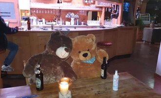 Zwei große Stoffbären sitzen an einem Tisch in einer Bar. Auf dem Tisch stehen zwei Bierflaschen und eine Kerze.