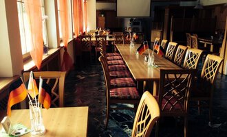 Ein einladendes Restaurant mit langen Holztischen und Stühlen. An den Tischen stehen kleine deutsche Fahnen und Fenster mit roten Vorhängen sorgen für eine gemütliche Atmosphäre.