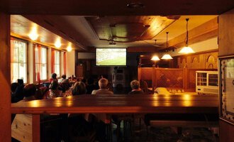 Eine Gruppe von Menschen sitzt in einem gemütlichen Raum und schaut ein Fußballspiel auf einem großen Bildschirm. Die Atmosphäre ist entspannt und gesellig.