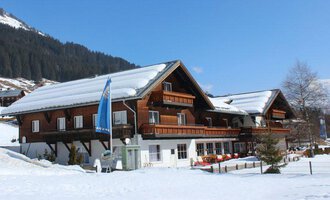 Ein traditionelles Holzhaus im Schnee, umgeben von Bergen. Der Himmel ist klar und blau, und es gibt viele schneebedeckte Flächen.
