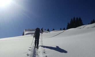 Ein Wanderer erklimmt einen schneebedeckten Hang unter einem klaren blauen Himmel. Sonnenlicht reflektiert das weiße Schnee, während der Weg sichtbar ist.