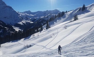 Ein Skifahrer steht auf einer schneebedeckten Landschaft mit hohen Bergen im Hintergrund. Der Himmel ist klar und blauer Sonnenschein strahlt auf die Szene.