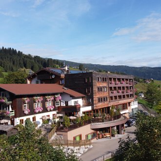 Ein großes Hotelgebäude in ländlicher Umgebung mit grünen Hügeln im Hintergrund. Der Himmel ist klar und die Landschaft wirkt einladend. | © Das Alphotel Hirschegg Hotel GmbH | Mark Kerwell