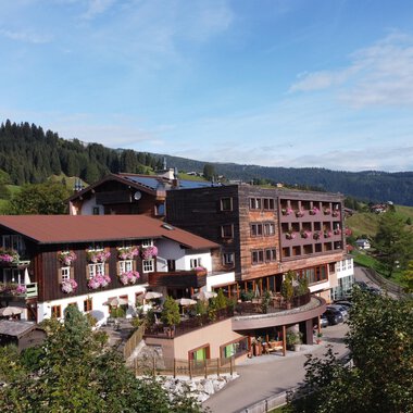 Ein großes Hotelgebäude in ländlicher Umgebung mit grünen Hügeln im Hintergrund. Der Himmel ist klar und die Landschaft wirkt einladend. | © Das Alphotel Hirschegg Hotel GmbH | Mark Kerwell