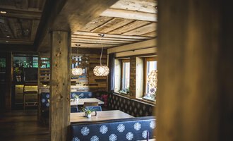 Eine gemütliche Inneneinrichtung mit Holzbalken und einem stilvollen, modernen Design. Am Tisch stehen Pflanzen und es gibt auffällige Lampen sowie Fenster mit natürlichem Licht. | © Das Alphotel Hirschegg Hotel GmbH | Dominik Berchtold