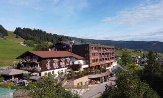 A large hotel building in a rural setting with green hills in the background. The sky is clear and the landscape looks inviting. | © Das Alphotel Hirschegg Hotel GmbH | Mark Kerwell