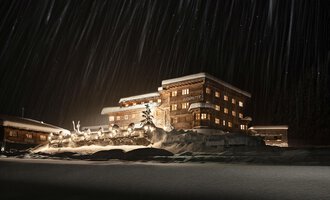 Ein gemütliches Holzgebäude im Schnee, beleuchtet von warmem Licht. Im Hintergrund fallen sanft Schneeflocken durch die Nacht. | © Auenhütte | Yorck Dertinger