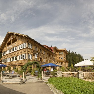 Ein rustikales Hotel mit Holzfassade in einer schönen Naturlandschaft. Der Außenbereich hat Sitzgelegenheiten und Sonnenschirme unter einem bewölkten Himmel. | © Auenhütte | Yorck Dertinger