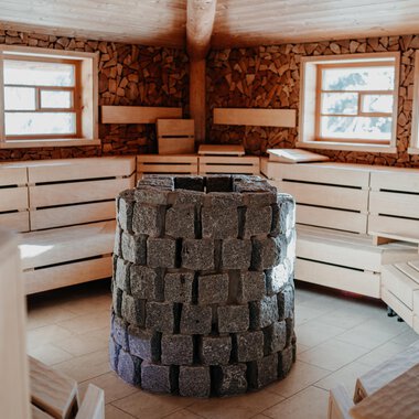 Ein gemütlicher Saunaraum mit Holzbänken und großen Fenstern. In der Mitte steht ein Ofen aus Steinen. | © Verwöhn- & Wellnesshotel Walserhof| Aileen Melucci